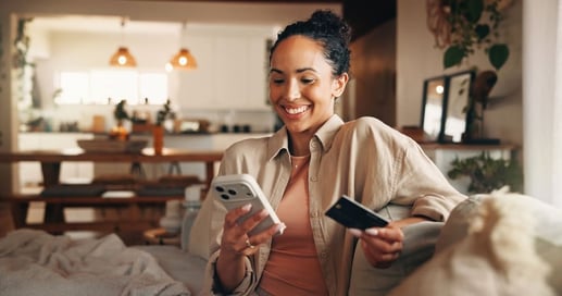 Mujer sonriente en su casa usando el celular y una tarjeta de crédito para realizar el pago total de su mes.