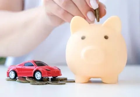 Mano depositando una moneda en una alcancía junto a un carro de juguete rojo sobre monedas: cuenta de ahorro programado para vehículo