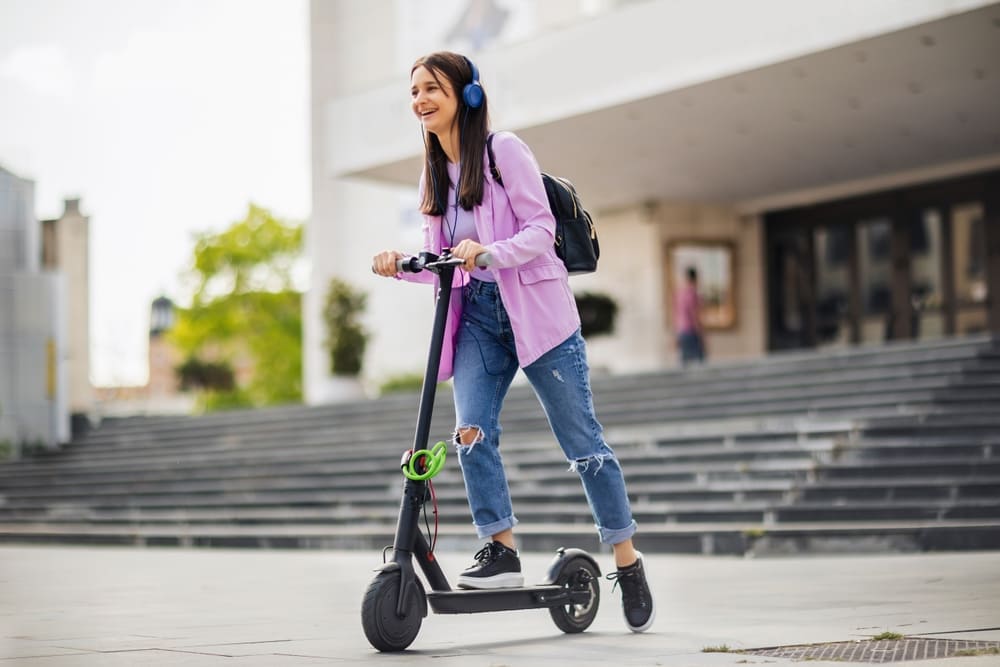 Estudiante universitario sonriente desplazándose en una patineta eléctrica por la ciudad.