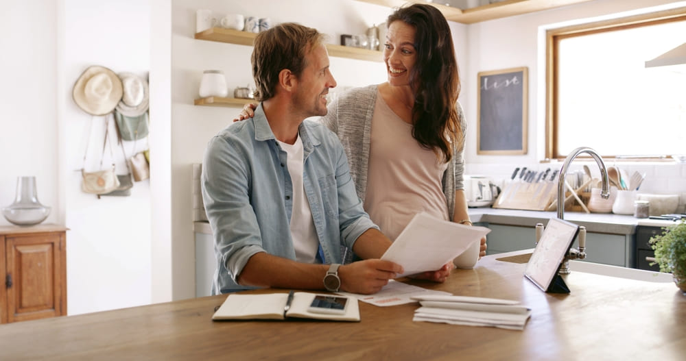 Pareja sonriente en la cocina revisando documentos y un certificado de cuenta de ahorros para un trámite de arrendamiento.