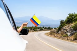 Mujer sonriente sosteniendo la bandera de Colombia desde la ventana de un vehículo durante un viaje por carretera en un puente festivo.