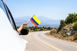 Mujer sonriente sosteniendo la bandera de Colombia desde la ventana de un vehículo durante un viaje por carretera en un puente festivo.